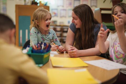 A small child smiling and drawing with her teacher sitting next to her