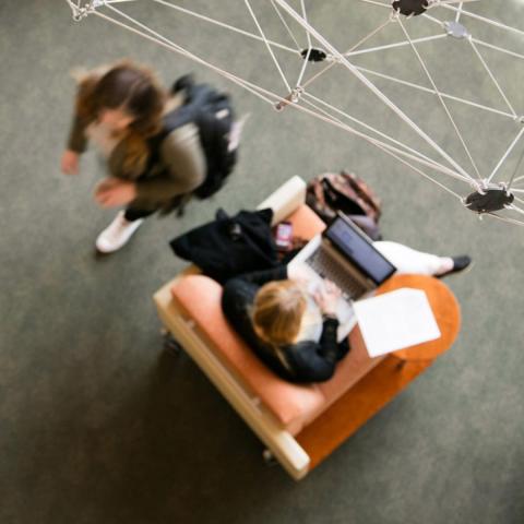 Overhead view of student sitting in chair working on laptop while another student walks by