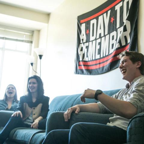 Students sitting on a couch in a dorm room laughing with "A Day to Remember" banner hanging on the wall