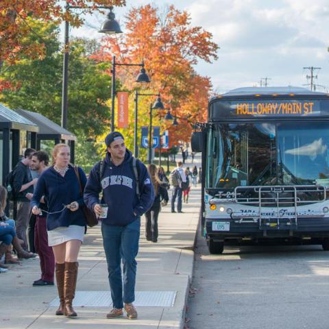 Picture of a UNH bus at the bus stop on Main St. with students walking by on the sidewalk